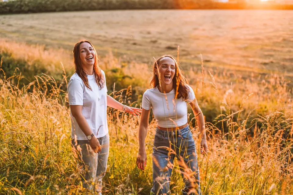 Twee zussen wandelen lachend door het hoge gras in het avondlicht
